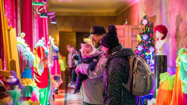 Parents with baby standing in long gallery at Mottisfont, decorated with acid-coloured fabrics and pantomime costumes, for Christmas